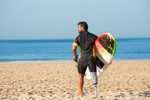 Back view full body muscular male surfer with leg prosthesis standing with surfboard on sandy sunny seashore and looking away
