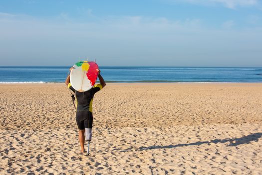 一名带假肢的冲浪者拿着冲浪板，走在阳光明媚的葡萄牙海滩上