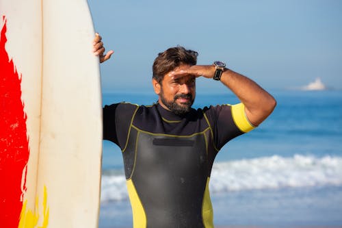 Handsome fit ethnic male surfer in wetsuit standing near surfboard on seashore and looking away while covering eyes from sun by hand