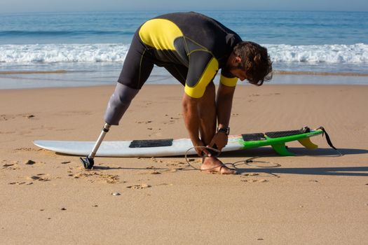 Full length sporty tanned male surfer with leg prosthesis tying surfboard leash around ankle while standing on sandy seashore before surfing