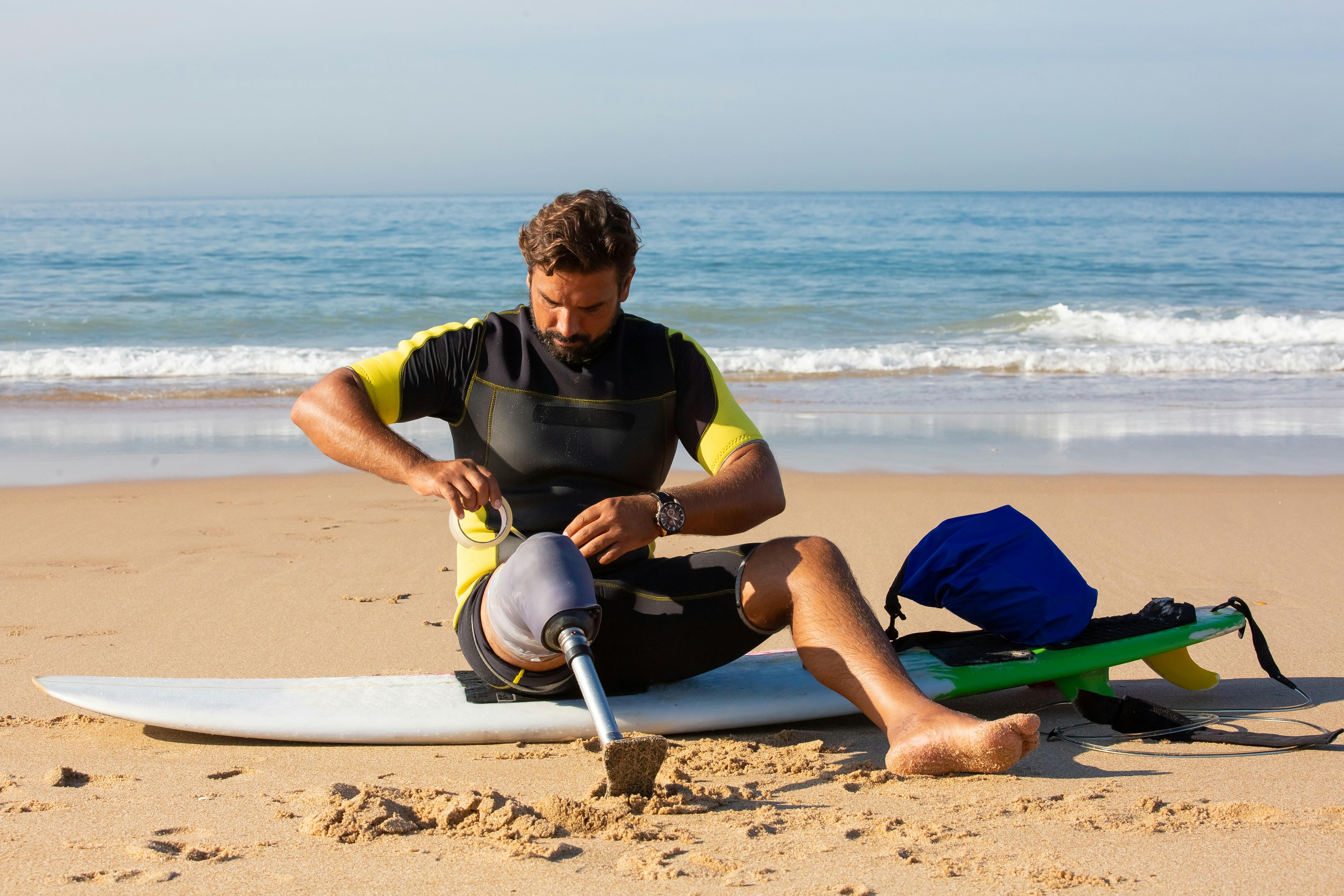 concentrated amputee male surfer wrapping prosthesis with tape on beach