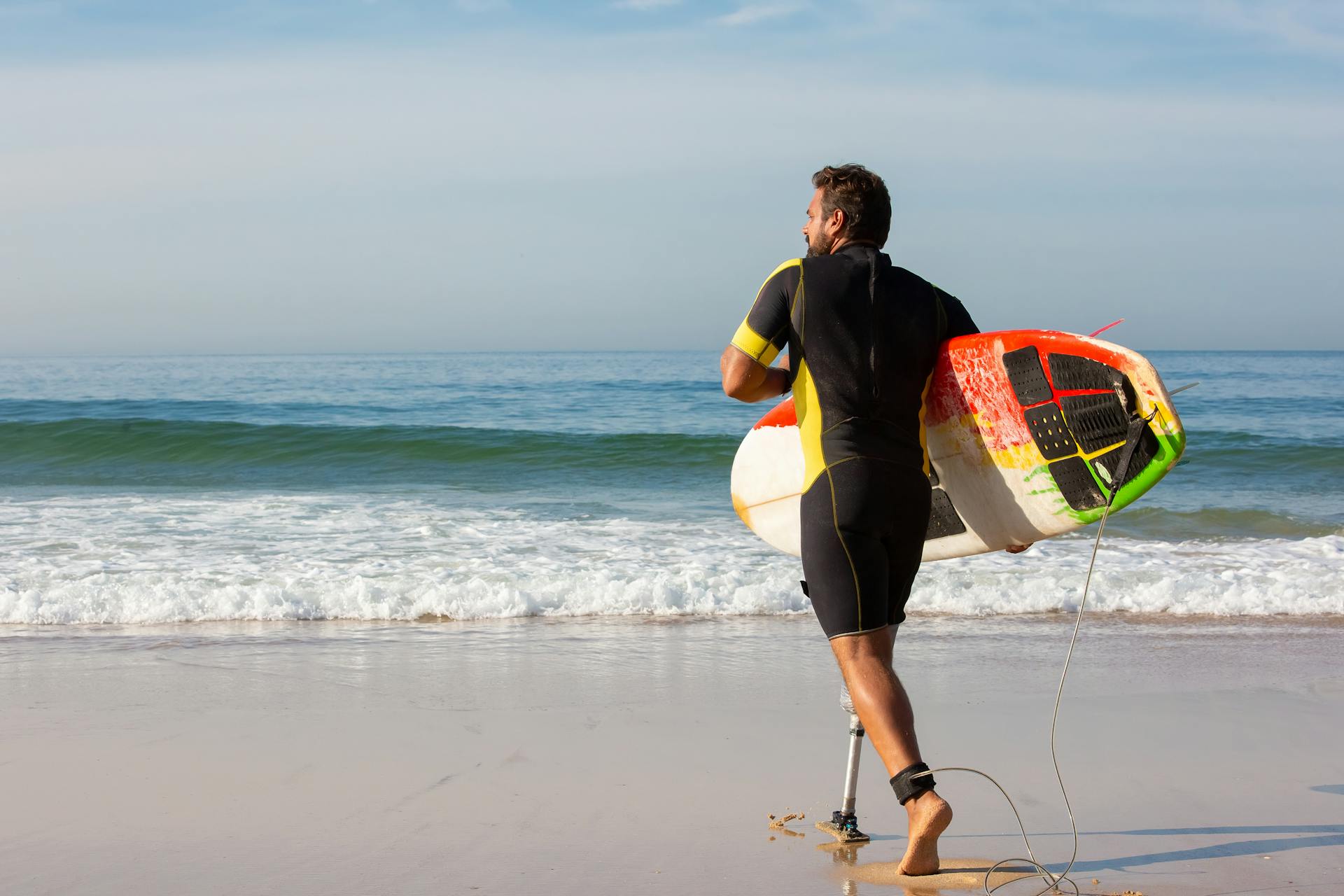 Back view unrecognizable energetic male surfer with leg prosthesis carrying surfboard and walking on wet sandy coast towards foamy sea