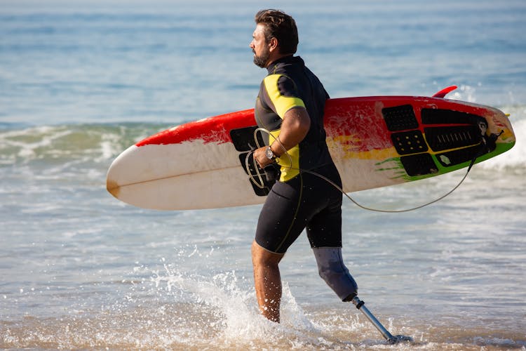 Sporty Male Surfer Amputee With Surfboard Running In Shallow Seawater