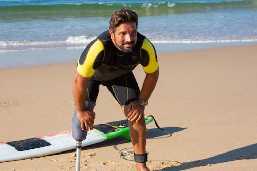 Free Tired ethnic male surfer in wetsuit standing in recovery position with hands on knees on sandy seashore near surfboard and looking away Stock Photo