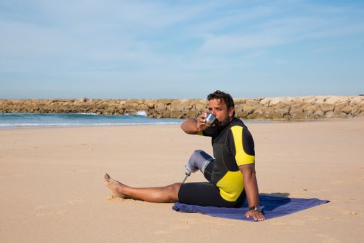 Side view full body tanned ethnic man with amputated leg wearing wetsuit resting on towel on sandy beach and enjoying hot drink from thermos cup while looking at camera