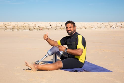 Side view full length handsome ethnic man with leg prosthesis resting on sandy seashore and showing thumb up gesture while looking at camera with smile