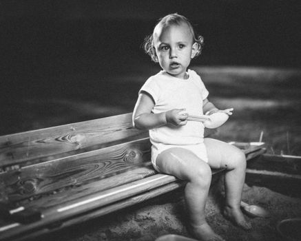 Charming black and white image of a toddler sitting on a bench outdoors.