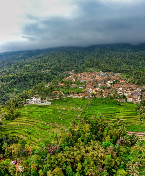 Free Majestic drone view of small settlement located on hill slope near terraced rice paddy fields and lush green rainforest against cloudy sky Stock Photo