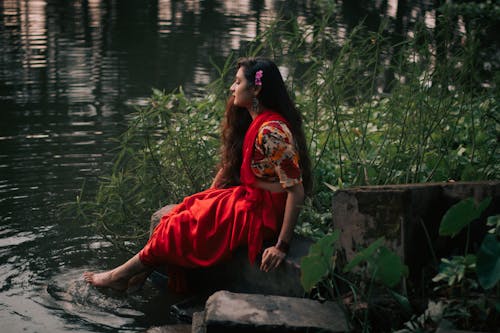 Woman in Red Traditional Dress Soaking His Feet on the Lake while Sitting on a Big Rock