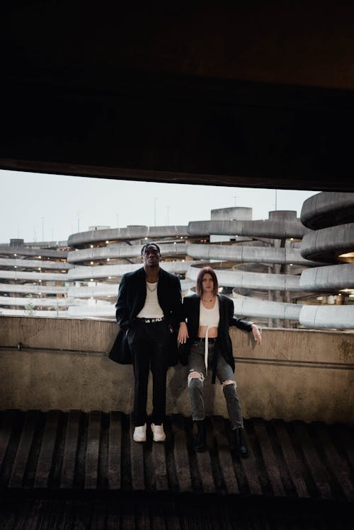 Free Full body of young confident woman and black man in trendy outfits leaning on concrete wall in multistory parking garage against urban background Stock Photo
