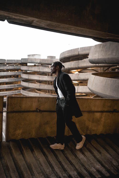 Side view of young black male in trendy clothes and sneakers walking with hands in pockets on parking ramp in urban building and looking out balcony