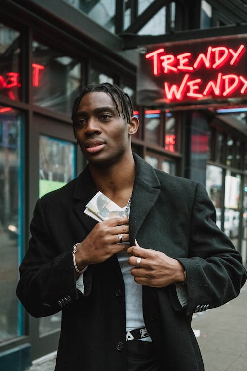 Self assured young African American guy with dreadlocks in trendy outfit putting folded newspaper in coat pocket while standing on city street