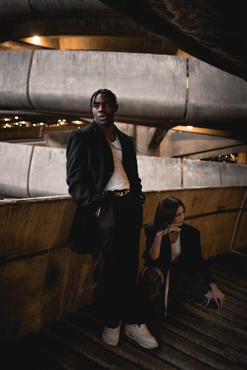 Confident young African American guy in stylish clothes standing with hands in pockets near trendy woman sitting on haunches in modern building