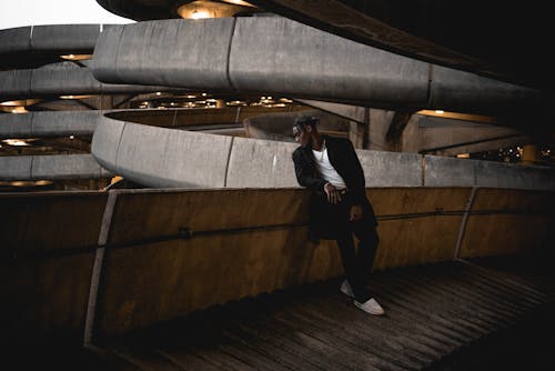 Full body of young stylish African American male in casual clothes looking out balcony of multistory parking garage against urban background