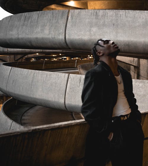 Thoughtful young black guy with dreadlocks in trendy outfit chilling on balcony of modern building with hands in pockets