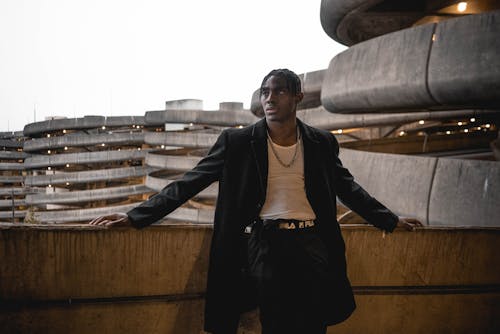 Serious young African American man with dreadlocks in trendy outfit standing in multilevel parking lot and looking away against urban background