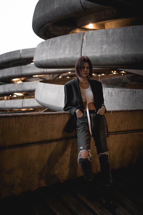 Full body of confident young female in trendy crop top and ripped jeans standing on ramp of multistory parking building and looking at camera against urban background