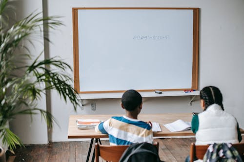 Free Back view unrecognizable multiethnic pupils sitting at desk with copybooks against whiteboard in light modern classroom Stock Photo