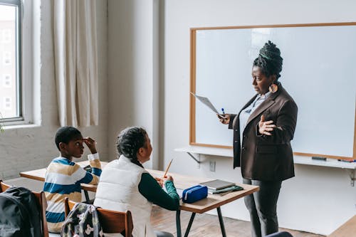 Adult African American female teacher in formal suit standing near whiteboard and explaining new material to multiethnic elementary pupils in light classroom