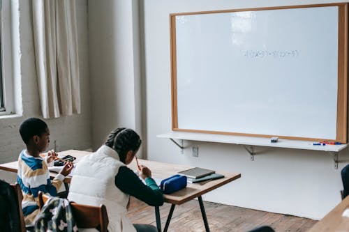 Free Back view African American elementary pupils in casual wear sitting at desk in light classroom and doing assignment Stock Photo