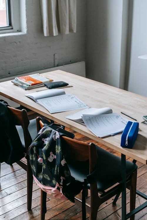 Wooden desk with opened copybooks and workbooks placed near chairs with backpacks in light classroom