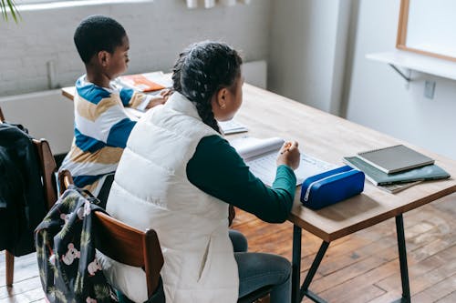 Free Back view focused elementary ethnic pupils in casual clothes sitting at desk with workbooks while attending class Stock Photo