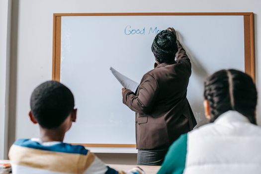 Back view unrecognizable African American female teacher wearing formal clothes writing on whiteboard for multiethnic elementary classmates