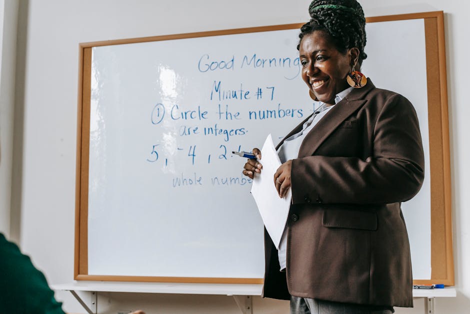 Crop smiling African American female teacher standing near whiteboard with papers in hands during math lesson