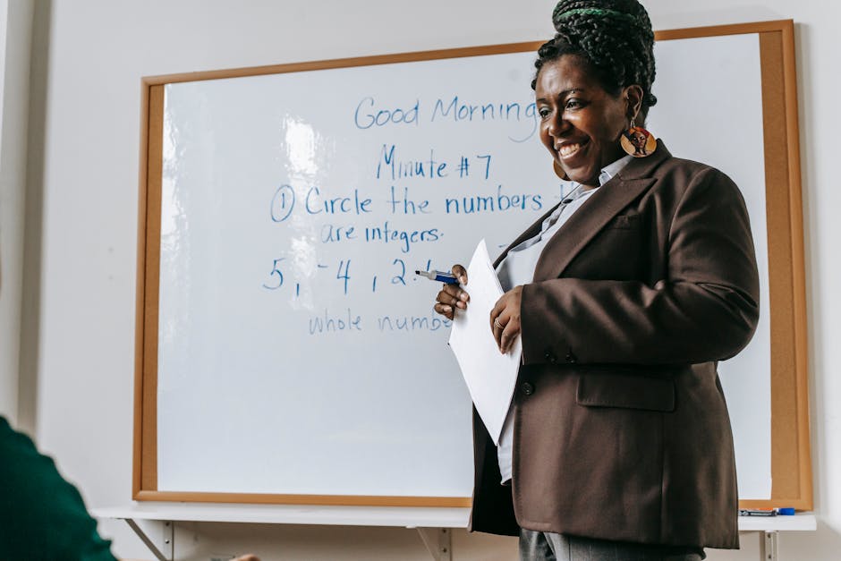 Crop smiling African American female teacher standing near whiteboard with papers in hands during math lesson