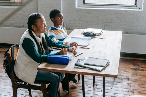 Free Side view glad multiethnic elementary pupils sitting at school desk with notebooks and textbook and looking away Stock Photo