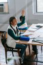 Side view full body content Asian schoolgirl in casual clothes sitting at desk with stationery and raising hand