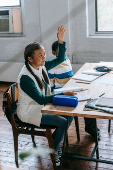 Side view full body content Asian schoolgirl in casual clothes sitting at desk with stationery and raising hand