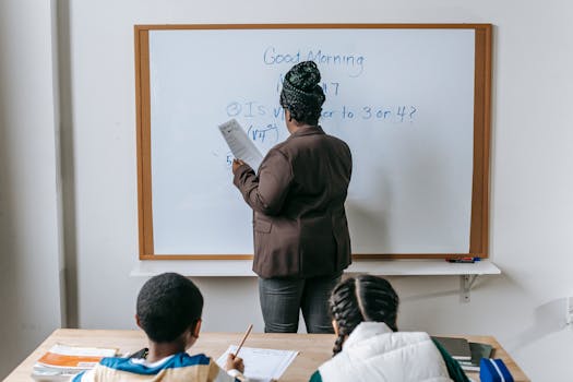 Teacher writing math lesson on whiteboard while students take notes in a classroom setting.
