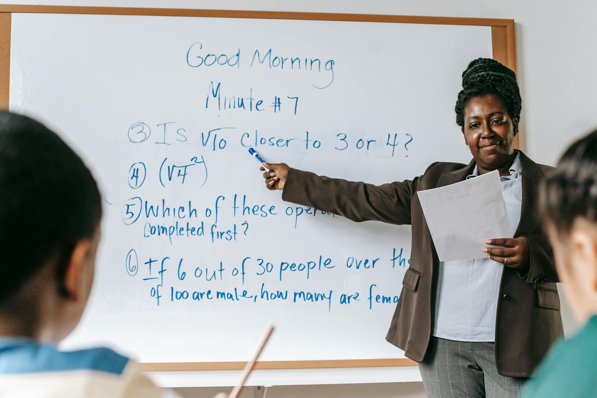 Teacher explaining math problems on whiteboard in a lively classroom setting.