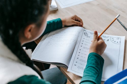 High angle crop unrecognizable schoolgirl sitting at desk with stationery and doing homework