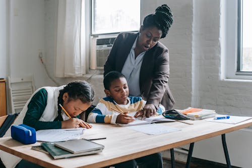 Free Focused African American female teacher in formal clothes standing near school desk with diverse pupils and pointing at schoolboys copybook Stock Photo