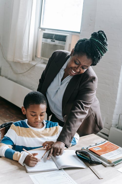 High angle of positive adult African American female teacher in formal clothes helping little boy doing assignment during lesson in school