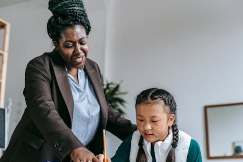 Middle aged African American female teacher with dreadlocks in formal clothes helping smart attentive little Asian girl solving exercise during lesson