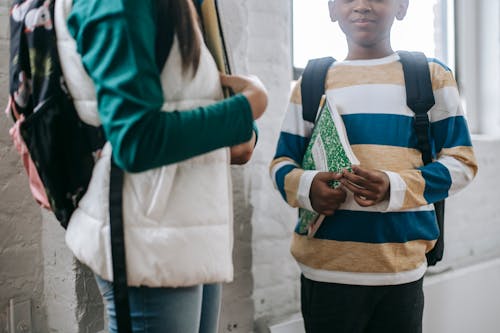 Free Crop anonymous multiethnic little friends in casual clothes and backpacks standing in school with notebooks and speaking during break Stock Photo