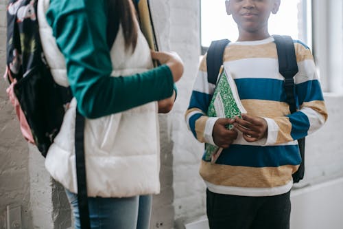 Crop anonymous multiethnic little friends in casual clothes and backpacks standing in school with notebooks and speaking during break