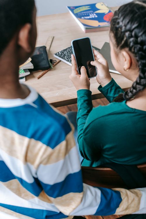 Free Back view of crop unrecognizable diverse friends surfing internet on smartphone while sitting together at wooden desk in classroom during break Stock Photo
