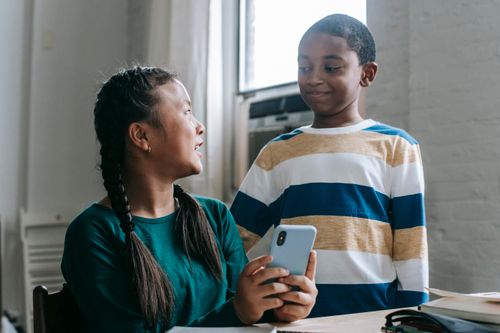 Free Little Asian schoolgirl sitting at table in classroom and sharing mobile phone with smiling African American friend during break Stock Photo