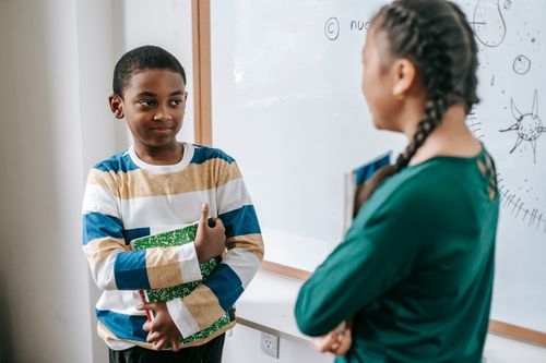 Free Smart African American schoolboy standing near whiteboard with stack of textbook and answering question with anonymous ethnic classmate during lesson Stock Photo