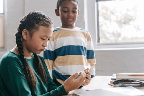 Funny little Asian girl playing game on smartphone while spending break with black classmate at school