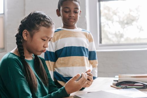 Free Funny little Asian girl playing game on smartphone while spending break with black classmate at school Stock Photo