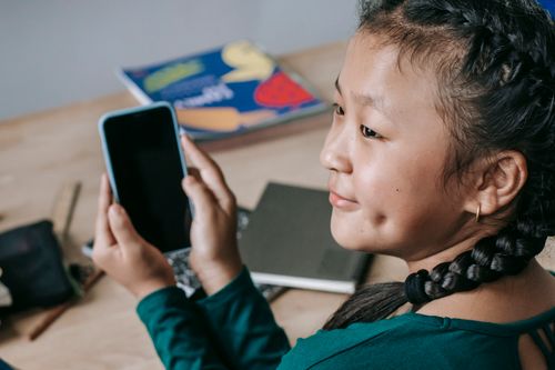 Free From above of crop adorable little Asian girl with braids sitting at desk in classroom and browsing smartphone during break Stock Photo