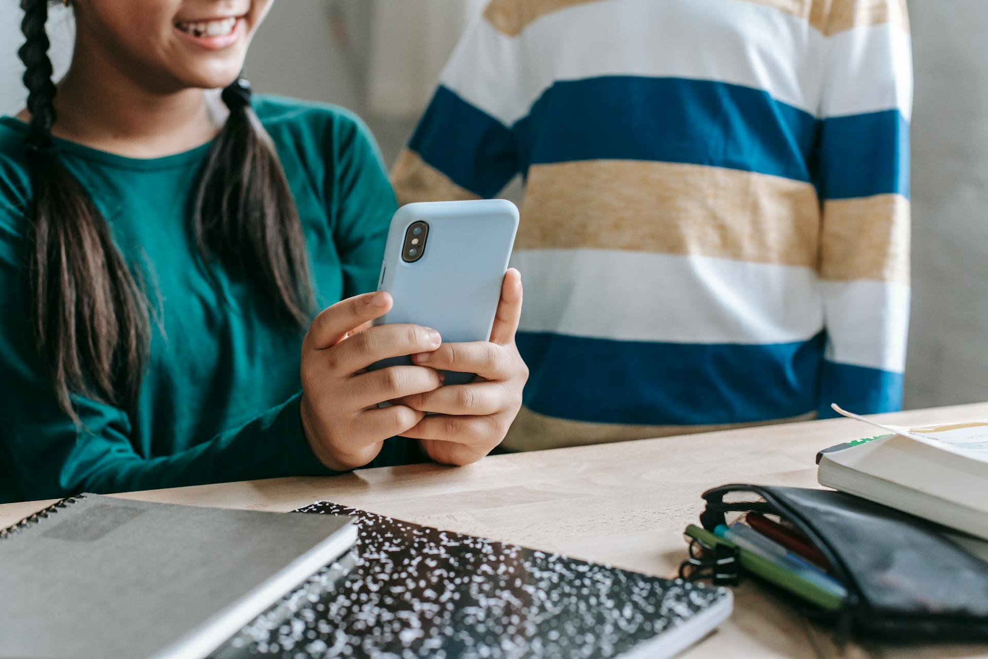 Crop anonymous cheerful little schoolgirl in casual clothes sitting at table in classroom and sharing video on smartphone with unrecognizable friend