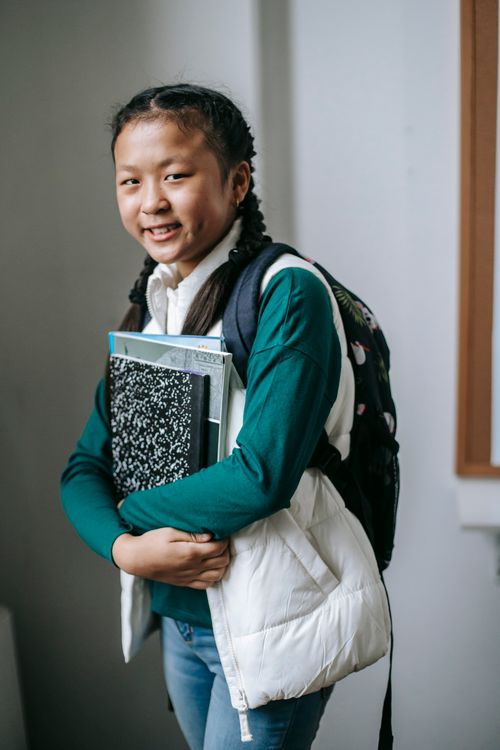 Free Side view of positive little Asian girl in casual clothes and backpack smiling and looking at camera while standing in classroom with pile of books Stock Photo