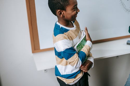 Free Side view of crop happy little African American schoolboy standing near whiteboard with stack of textbooks and smiling Stock Photo