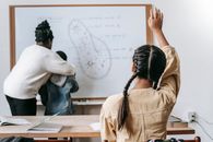 Back view of faceless girl with pigtails sitting at desk and raising hand while teacher  with boy standing near whiteboard and writing